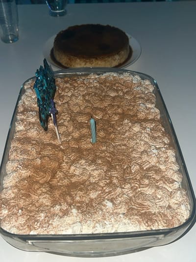 A rectangular glass dish filled with tiramisu topped with cocoa powder, a blue and silver spaceship decoration, and a blue birthday candle. A flan sits on a plate next to the tiramisu. Two glass cups are partially visible in the foreground. The image is taken from an overhead perspective, slightly angled.