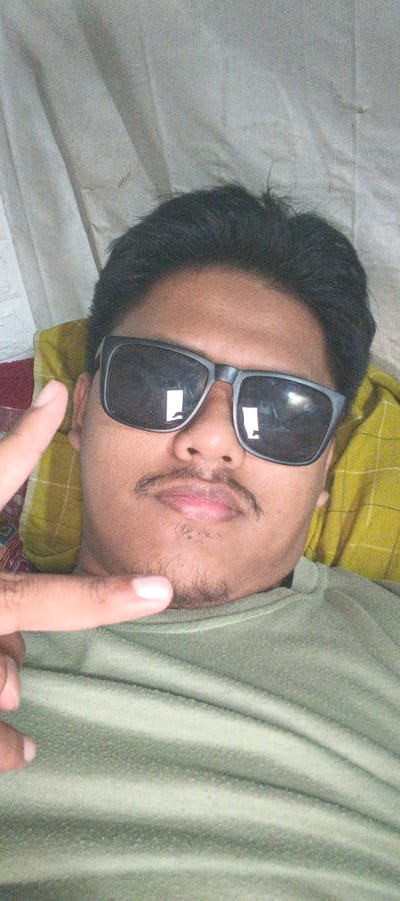A selfie of a person is taken from a high angle, looking down at the camera. The person is lying down on a yellow pillow with a white and gray patterned blanket behind them. The person is wearing a green t-shirt and black sunglasses. Their hair is dark and short. They have a mustache and a small beard. The person is holding up their left hand with two fingers extended in a peace sign. The background is a white cloth with some folds and creases. The image is slightly grainy and the lighting is dim. It is not possible to determine the location in the city of Kemuning, Indonesia.