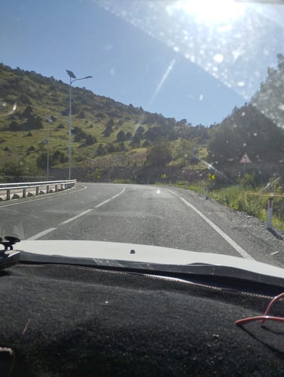 The image shows a road with guardrails and solar-powered streetlights on a hillside. There are road signs indicating curves. The scene appears to be a scenic route, possibly in a rural or mountainous area, with sparse vegetation. The road is asphalt, with white lane markings. The sky is clear and blue, with the sun high in the sky, creating some glare on the windshield. The foreground shows part of a car's dashboard. Based on the provided location of Myk, Uzbekistan, this scene is likely a road leading into or out of the city, showcasing its natural landscape.