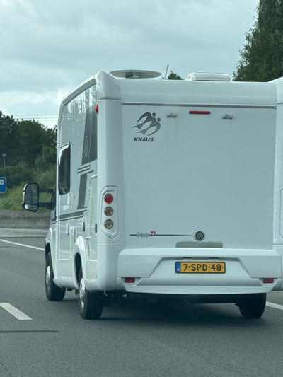 A white KNAUS motorhome with license plate 7-SPD-48 is driving on a highway. The motorhome is white with black and grey decals. It has a mirror on the driver's side and a blue sign with white text and a white square on a pole is visible on the left side of the road. The sky is cloudy and the road is asphalt. The image appears to be taken from another vehicle. The specific location in Meppel, Netherlands is not identifiable from the image.