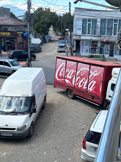 The image depicts a street scene in Khashuri, Georgia, with several vehicles and buildings visible. In the foreground, a white Ford Transit van is parked on a gravel road, its license plate reading "OV-764-OV". To the right of the van, a red Coca-Cola truck is parked, its side emblazoned with the iconic Coca-Cola logo. Adjacent to the truck, a white cab of another truck is visible, with the number "309" displayed on its side.
Further down the street, more vehicles are present, including a silver car and a green Lada hatchback. Buildings line the street, with one structure featuring a sign with "APOTEKA" written in large letters. Another building has signs with Georgian script. The street is unpaved and appears to be in a somewhat neglected state. Overhead, a complex network of electrical wires crisscrosses the sky. The overall impression is a bustling, everyday scene in a Georgian town.