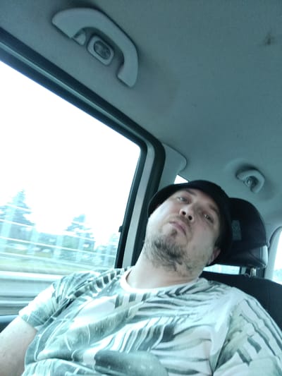 The media file is a selfie taken by a man inside a vehicle. He is wearing a black hat and a t-shirt with a tropical leaf pattern. The man has a beard and appears to be resting his head against the headrest. The background shows trees and a road, suggesting the vehicle is in motion. The lighting is bright, possibly indicating daytime, and the sky is overcast. The interior of the car is visible, including a grab handle and part of the ceiling. The overall mood appears relaxed or tired. The location is Průhonice, Czechia. There is no visible text in the image.