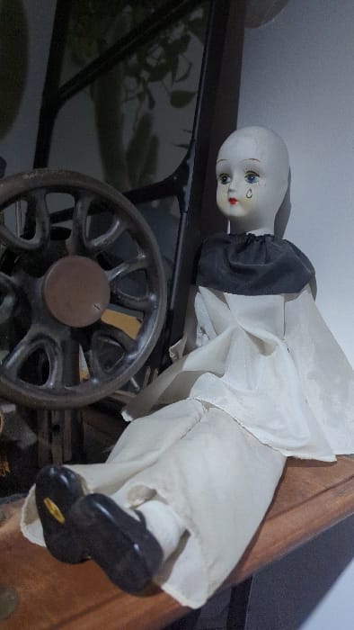 An antique-style doll, likely made of porcelain or bisque, is depicted in an indoor setting. The doll's head is the primary focus, featuring delicate painted blue eyes, red lips, and a distinct blue teardrop painted beneath its left eye. It appears to be bald or has its head covered. The doll is dressed in a flowing, off-white or cream-colored garment with large sleeves that gather at the cuffs in black fabric. Small black shoes are visible on its feet.
The doll is positioned on a dark, flat surface, likely a piece of wooden furniture. In the foreground, a portion of a cast iron wheel with spokes is prominent, strongly resembling the flywheel or treadle wheel of an antique sewing machine. To the right, another piece of wooden furniture with a warm brown tone is visible. The background consists of a dark, reflective surface, which could be a window or a glass display case, showing blurred reflections of green plants and what appears to be an interior ceiling or wall. The lighting is soft and natural, suggesting it is daytime. The image captures a static display, possibly in a home or a collection, in Châtenois, France. There is no visible text.