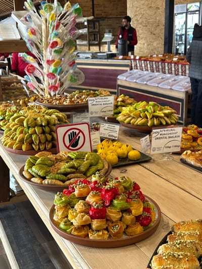 An indoor marketplace or shop in Tsovagyugh, Armenia, displays a wide variety of pastries and confections arranged on wooden counters. In the foreground, numerous dishes hold meticulously stacked sweets. A brown ceramic plate on the left showcases green, folded pastries. To its right, another brown plate presents a vibrant pyramid of small, cylindrical pastries in red, orange, yellow, and green hues, each topped with crushed pistachios. Further right, a black tray holds golden-brown pastries also dusted with pistachios. White price tags are interspersed among the displays, labeling items like "ORIENTAL BAKLAVA" and "DUBAI CHOCOLATE" with prices in Armenian Dram. A prominent white sign with a red border and a black "do not touch" symbol (an open hand with a diagonal line) is positioned centrally among the sweets. In the upper-middle, a wooden stand is laden with individually wrapped, colorful lollipops. Behind the main counter, a man with dark hair and a beard, wearing a red shirt and dark vest, stands near a weighing scale and other equipment. To his right, a person is partially visible from the waist down, wearing dark pants and a dark jacket. The background features light-colored wooden paneling on the walls and a long wooden counter with sections covered by clear, rectangular lids. The floor consists of light wooden planks.