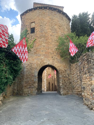 This media file shows an outdoor scene in San Quirico d'Orcia, Italy, with a focus on a stone tower and an arched entryway. Several red and white patterned flags are prominently displayed, some attached to poles extending from the stone structures, and others seen fluttering in the background. The tower itself is made of large, rough-hewn stones, with a small window and ivy growing on its side. The entryway is a dark archway, suggesting a passage into another part of the complex. The ground is paved with grey, diamond-shaped stones. The sky above is cloudy, indicating it might be daytime. The overall impression is of an historic, possibly medieval, setting decorated for a celebration or event, given the prevalence of flags. There is no visible text in the image.