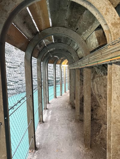The image depicts a man-made pedestrian walkway or tunnel in Pellafol, France, built along a natural rock face and overlooking a body of water. The pathway extends longitudinally into the distance.

On the left side of the concrete path, a protective barrier consisting of a vertical metal mesh fence and multiple horizontal wire ropes secures the walkway from an expansive body of vivid turquoise-blue water. Beyond the water, a grey rock wall, partially covered by a protective mesh, is visible.

On the right side, a series of vertical concrete pillars support the overhead structure and abut a rugged, natural rock wall. A bundle of utility cables, including grey, green, and orange conduits, runs horizontally along the upper section of this right rock wall, affixed to the structure.

The overhead structure comprises a repeating series of concrete arches, interspersed with sections of wooden planks and mesh, forming a semi-enclosed ceiling.

The pathway floor is composed of rough, light-colored concrete. Natural light illuminates the scene, entering primarily from the left over the water and from a brighter opening or light source at the distant end of the walkway. No individuals are present within the frame. The structure exhibits signs of weathering and appears utilitarian.