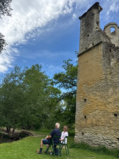 The image shows a tall stone structure, likely a tower or part of a castle ruin, in West Oxfordshire, United Kingdom. The structure is made of rough-hewn stones and shows signs of age and weathering. It rises high against a blue sky with scattered clouds. At the base of the structure, in a grassy area, two people are sitting on green chairs. One person is older with white hair, wearing a dark shirt and shorts. The other person, possibly a woman, is wearing a white top. They appear to be relaxing or enjoying the view of the stone structure amidst the surrounding greenery, including various trees and foliage. There is a stream with an exposed tree root system winding nearby.
