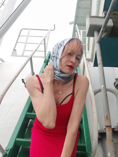 A woman with pink hair, wearing a red dress and a patterned headscarf, poses on a green metal staircase. She is looking at the camera with red lipstick and a necklace with a small pendant. The staircase appears to be on a boat or ship, with metal railings and white surfaces visible in the background. The overall lighting suggests it is daytime, with overcast weather. The woman's expression is direct and confident. There is no visible text in the image.