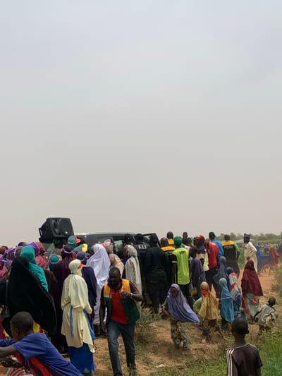 The image depicts a crowd of people gathered around a black SUV and a large speaker in a rural area of Ringimawa, Nigeria. The coordinates provided (11.667, 8.416) indicate a location in the northeastern part of the country. Several men and women are standing in the crowd, some of whom are wearing brightly colored clothing or traditional Islamic garb. In the foreground, a man in a red shirt is walking away from the group while a young boy in a black and white striped shirt watches. The scene is set against a backdrop of dry, brown grass and a dusty sky.