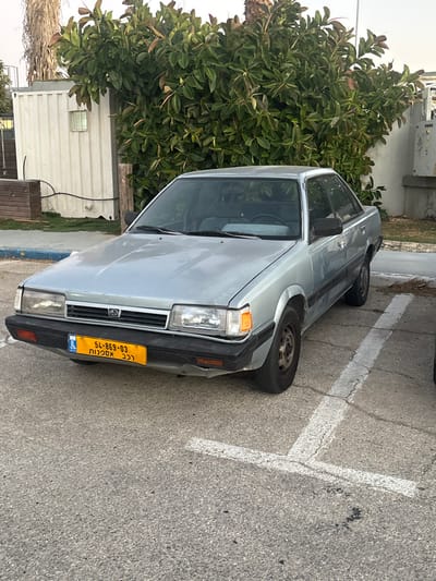 The image displays a light blue/silver four-door Subaru sedan, an older model, parked in a paved lot in Mazkeret Batya, Israel. The vehicle is positioned within a marked parking space, defined by faded and cracked white lines on an asphalt surface. The front of the car is visible, featuring rectangular headlights, amber turn signals, a chrome grille with a central Subaru logo, and a black bumper showing minor damage on its lower left side. An Israeli yellow license plate, displaying the number "54-869-03" in black, is mounted on the front bumper. Below the number, the Hebrew text "רכב אספנות" (Collector's Vehicle) is visible. The car's steel wheels are present, and the vehicle's paint shows some wear consistent with age.
In the background, a large, dense green bush or tree with broad leaves and some reddish accents is situated behind the car, extending across the center and right side of the frame. To the far left, a section of a palm tree trunk is visible. A white corrugated metal structure, resembling a utility shed or shipping container with a small window, is located behind the car on the left side, with a black cable running along its base. A light-colored building is partially visible on the far right. The scene is illuminated by natural daylight, with a bright, possibly overcast, sky overhead. No individuals or specific actions are depicted.