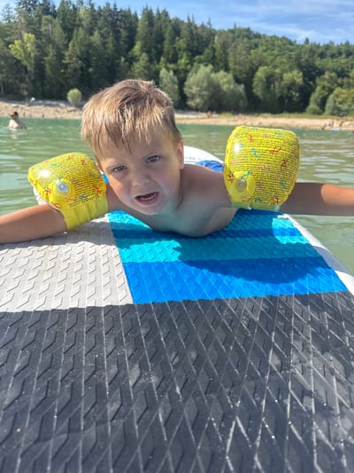 In Maisod, France, a young boy with blond hair and blue eyes is lying on his stomach on a paddleboard in a lake. He is wearing yellow arm floaties. The paddleboard is a light blue, white, and black color. In the background, there is a beach and a dense forest of green trees. The sky is clear and blue.