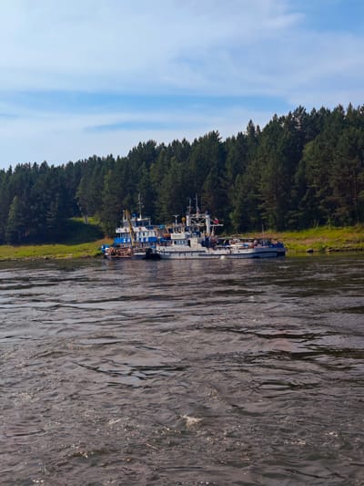 In Porog, Russia, a calm river flows in the foreground, with a dense forest of evergreen trees lining the opposite bank. Moored along the shoreline are two barges and a smaller boat. The barges appear to be industrial, with cranes and cargo visible. The sky above is partly cloudy, with a few wispy clouds scattered across a pale blue expanse. The overall scene suggests a quiet, working waterfront in a natural setting.