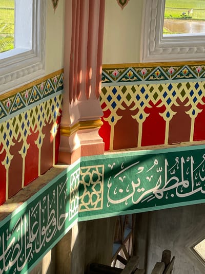 This image depicts the brightly lit interior of a building in Mns Asan, Indonesia, offering a view of the exterior through a window. The interior walls are richly decorated with various patterns and colors.
The upper wall section is painted a pale yellow. Below this, a horizontal decorative band features several layers: a gold stripe, a light teal band with pink and yellow floral motifs, a gold band with a dark blue and gold diamond pattern, and a prominent red section adorned with brown, stylized arch-like shapes and blue teardrop details. A fluted, pale pink column with a gold-painted base is situated in the corner, illuminated by sunlight and casting shadows.
Below this decorative band, a green balustrade or railing features intricate white Arabic calligraphy and a gold octagonal geometric pattern. Sunlight casts diagonal shadows across this green surface.
Through a white-framed window on the left, an exterior scene is visible: lush green rice paddies, partially flooded with water. In the distance within the paddy field, a single individual, presumably a farmer, is visible wearing a white hat and working in the field. A small red object or marker is near this person. The area beneath the green railing appears to be a lower level or an unfinished section, with some wooden elements partially visible. No people are observed inside the building.