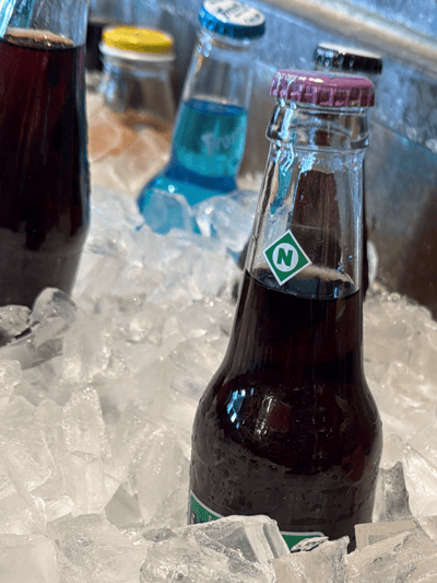 The image shows several glass bottles of different colored sodas submerged in ice. The soda on the left is a dark, deep red color. To its right, there is a soda with a yellow cap, and a blue soda with a blue bottle cap. In the right of the image, there is a bottle with a pink cap with a logo of the letter "N" inside a green rhombus. The ice around the bottles consists of relatively large, irregularly shaped pieces. The background appears to be a stainless steel surface, likely a cooler or container holding the ice and drinks. The image was taken in Rochester, United States.
