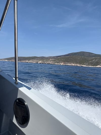 A boat is cutting through the deep blue Adriatic Sea, generating a white spray of foam along its starboard side. In the distance, a green, hilly island with rocky coastal cliffs is visible under a clear blue sky. A metal railing and part of the boat's white hull are in the foreground. The location is near Podhumlje, Croatia.