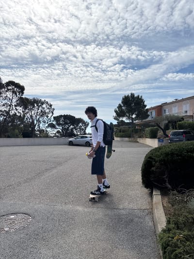 A photograph shows a young man skateboarding on a paved street in Fréjus, France. The individual wears a white long-sleeved shirt, jean shorts, and black and white athletic shoes with white socks. He carries a backpack with a skateboard attached to it. In the background, there are houses, trees, parked cars, and a partly cloudy sky. A street sign reading "Oradour" is visible near one of the buildings. A manhole cover is visible on the street.