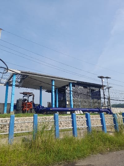 The image shows a water pump station located at the coordinates -7.522, 112.705 in Kecamatan Gempol, Indonesia. The station is protected by a fence with blue railings, and a large diesel engine is visible behind the fence. There is a large metal pipe attached to the engine, and it is connected to a smaller pipe that runs alongside the fence. The engine is partially obscured by a structure with a metal roof. Multiple power lines cross over the scene. There are no people or animals visible in the image. The sky is clear and blue. The ground is covered in green grass, which appears to be growing in a wild and unkempt fashion.