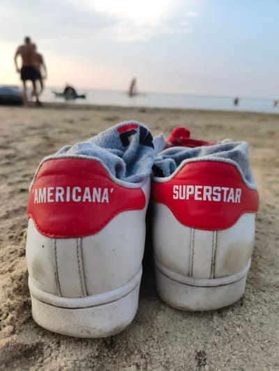 The image shows the back view of two white Adidas sneakers with red accents on a sandy beach. The left shoe has the text "'AMERICANA'" printed in white on the red heel tab, and the right shoe has the text "SUPERSTAR" printed in white on its red heel tab. Light grey socks, with some black and red details, are visible inside both shoes. In the blurred background, several people are visible near the water, and a windsurfing sail can be seen on the horizon. The environment is a beach with sand in the foreground and the ocean and sky in the background. The lighting suggests it is late afternoon or early evening. The location is not specified as Gubernskoe, Russia.