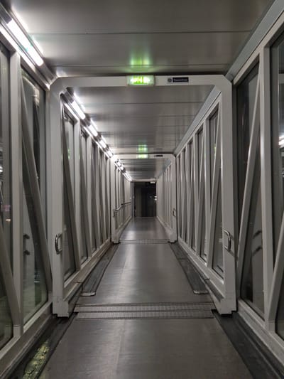 The image depicts an interior view looking upwards inside what appears to be a fire escape tunnel or passageway. The walls are metallic, with vertical seams suggesting panels. On the left wall, there's a ThyssenKrupp logo and an illuminated green exit sign. The ceiling is constructed with a series of white, rectangular frames with supporting beams. Some of the frames seem to contain glass. The space narrows as it extends into the distance, with a small opening visible at the far end. There are no people visible. The lighting appears artificial and uneven, creating a play of light and shadow along the walls and structures. The specific location within Rionegro, Colombia, is not discernible from the image.