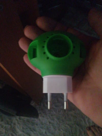 A close-up, dimly lit shot shows a person's hand holding a green and white object that resembles an electric mosquito repellent. The object has a green, bulbous top with small black dots and a white rectangular base with two metal prongs extending downwards. The background is dark and out of focus, with hints of a patterned surface that could be carpet or fabric. No specific location in the city of Степной, Russia is discernible from the image.