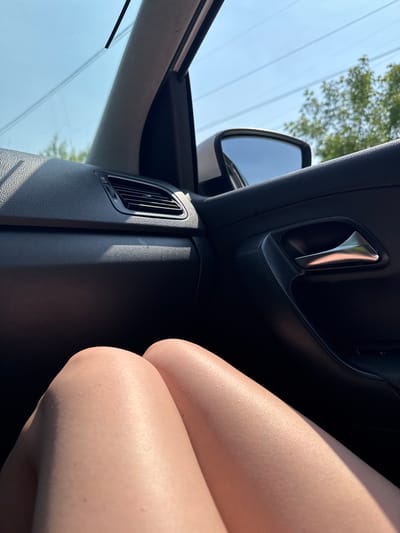 The image shows the interior of a car, with a person's legs in the foreground. The legs are light-skinned and appear to be bare. The interior of the car is dark, with a black dashboard and door panel. A silver door handle is visible on the right side of the frame. Outside the car, the sky is blue with some trees visible. The image does not contain any information or context related to the city of Leninsk-Kuznetsky, Russia.
