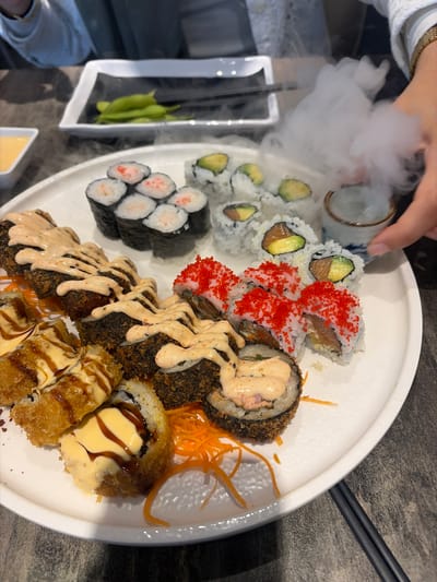 A white plate of assorted sushi, some of which are smoking, sits on a dark grey table. To the left of the plate, a small white dish of yellow sauce is visible. In the background, a black rectangular plate holds a serving of edamame beans. A hand with a gold bracelet is reaching for a small ceramic cup filled with a liquid, from which the smoke emanates. The setting appears to be a restaurant in Longueuil, Canada.