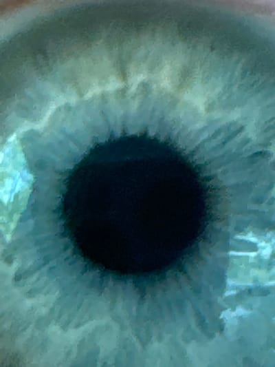 The image is a close-up of an eye, specifically the iris and pupil. The iris appears to be a light bluish-green color. The pupil is a dark black circle in the center. The texture of the iris is visible with a slightly rough appearance, possibly a stylized or painterly effect.
The location, city of Balakhna, Russia, cannot be confirmed from this image.