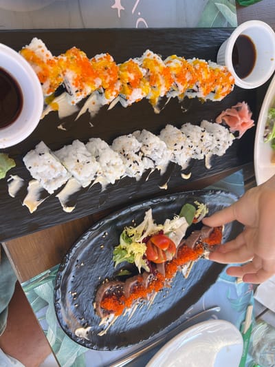 Two platters of sushi are presented on a wooden table, likely outdoors given the natural lighting. The left platter features a long row of maki rolls topped with orange tobiko (flying fish roe) and a creamy sauce. To the right of the maki, a smaller portion of sushi rolls is visible, sprinkled with black and white sesame seeds. A small white dish of soy sauce sits to the left of the maki, and a dollop of green wasabi is at the bottom left.
The right platter holds slices of seared tuna, also topped with orange tobiko and sesame seeds, accompanied by a small salad of lettuce and cherry tomatoes. A hand is reaching to pick up a piece of tuna from this platter, suggesting someone is about to eat. The setting appears to be a restaurant or cafe, with a tropical-themed placemat visible under the platters. White plates and cutlery are also present, hinting at a meal in progress. The time of day appears to be daytime, likely lunch or early dinner. The overall scene evokes a relaxed dining experience. No visible text is present in the image.
