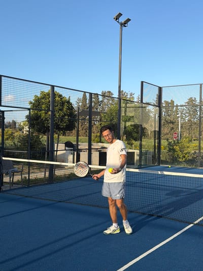 A man stands on a blue paddle tennis court, holding a paddle tennis racket in his right hand. He is wearing a white t-shirt and light gray shorts, with athletic shoes. Behind him is the court netting and wire fencing. The backdrop includes trees, foliage, and a "Stop" sign. The sky is clear and blue. A tall light post with two lights is to the left. The scene is in Tserkezoi, Cyprus.