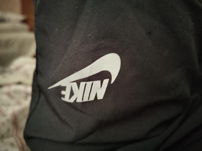 This is a close-up, horizontal shot of a black fabric with a white Nike logo. The logo is partially visible, with the swoosh and the letters "NIKE" in white font. The fabric appears to be made of a synthetic material, possibly polyester or nylon, and it has a slightly textured surface. There is a small dark mark on the fabric to the right of the logo. The background is out of focus and appears to be a blurred image of furniture and bedding with a floral pattern, suggesting the item might be clothing or a bag placed on a bed or sofa. The lighting is artificial and casts some shadows on the fabric, indicating an indoor setting, possibly a home. There are no people visible in the image. The specific location and time of day are not discernible from the visual cues. The primary subject is the Nike logo on the black fabric.
