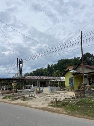 This is a vertical, eye-level, medium shot of a street scene in Rantau Pauh, Indonesia. The sky is overcast with thick, grey clouds. A prominent utility pole with numerous wires crisscrosses the frame, extending upwards towards the cloudy sky. To the right of the pole, a row of houses and buildings is visible. The first building is a two-story structure, with the upper floor painted yellow and featuring a blue-paned window. The lower part of the building appears to be under construction or in disrepair, with exposed bricks and scaffolding. Adjacent to this is another building with a rusted corrugated metal roof and a white metal fence. Further down the street, a paved road runs along the right side of the image, bordered by a grassy embankment with some litter. Several more buildings with various roofing materials and architectural styles line the street, interspersed with greenery. A yellow sign with black text is visible on the utility pole, displaying "MAKAN LEZAT BAKSO" and "AQIS" with additional smaller text. The overall impression is a slightly run-down but lived-in urban environment, likely in the daytime given the diffused light. The weather appears to be humid and possibly about to rain. There are no people visible in the frame.