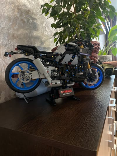 A detailed Lego Technic model of a Yamaha MT-10 SP motorcycle is displayed on a dark wooden surface. The motorcycle is black and gray with bright blue wheels. A Yamaha logo and "Rev Your Heart" are visible on a display plaque in front of the motorcycle. Behind the motorcycle, a large leafy green plant in a wicker basket is partially visible, as is a textured, light-colored wall. To the right of the plant, a small green toy vehicle and a hand are visible. The overall scene suggests an indoor display or collection. The location of "Берёзовая Роща, Russia" is not evident from the image itself.