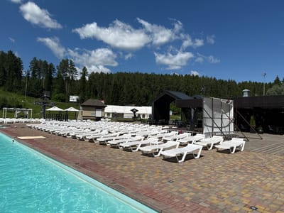 An aerial view from the east shows a pool and sunbathing area in Nechkino, Russia. The pool is on the left, and the sunbathing area is on the right with rows of white lounge chairs. The area is paved with interlocking bricks. Behind the sunbathing area, a large black tent is set up, possibly for an event. A forest is visible in the background. The sky is blue with some scattered white clouds.
