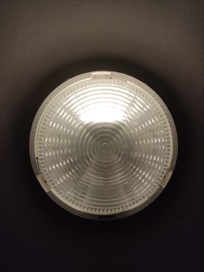 An overhead shot shows a circular ceiling light with a textured, clear plastic cover. The light inside is on and appears to be a fluorescent bulb, emitting a bright white glow. The cover has concentric rings and a bumpy texture, with a white plastic frame around the edge. The light is mounted on a dark, textured ceiling, with a shadow cast around it. The image is taken from a low angle, looking directly up at the fixture. The location is not specified in the image or its context, and therefore cannot be determined to be in Kolín, Czechia.
