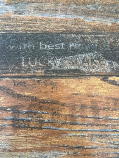 The image shows two wooden planks, one dark and the other lighter. The dark plank has light grey letters forming the words "with best luck". The lighter plank is brown and yellow, with grey marks that mimic wood grain. The planks are placed side-by-side and the texture is rough and worn.