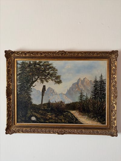 The image shows a framed oil painting of a mountain landscape. In the foreground, there is a dirt path winding through trees and bushes. A wooden signpost stands on the left side of the path. In the background, majestic snow-capped mountains rise against a pale blue sky with scattered clouds. The painting is displayed on a white, textured wall. There is no information available that connects this image or the artwork to the city of Osnabrück, Germany.