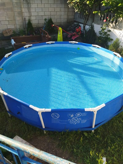 An elevated, wide-angle, outdoor shot captures a round, blue, above-ground swimming pool filled with clear water in Costești, Moldova. The pool's interior features a white grid pattern. The pool is surrounded by green grass and various objects. To the left, a rustic wooden boat is partially visible, with a white cloth draped over its edge. Behind the boat, a grey cinder block wall forms the backdrop, with a wooden pallet leaning against it. Further to the right, a red rototiller is parked near some evergreen shrubs and a tree. In the upper right corner, sunlight filters through the leaves of trees, casting shadows on a white wall adorned with ornate concrete trim and a pink rose in bloom. The pool's blue liner is supported by white PVC pipes and poles. Safety warnings are printed in multiple languages on the pool's exterior. In the foreground, a rusty, weathered blue metal railing runs horizontally across the bottom of the frame.