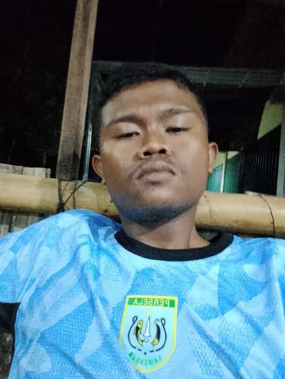 A close-up selfie taken at night shows a young man with dark skin and hair, looking directly at the camera. He is wearing a light blue t-shirt with a repeating abstract pattern and a yellow and green crest on the left chest. The background is dark and blurred, with hints of bamboo structures and some foliage visible. The setting appears to be outdoors, possibly a casual outdoor seating area. The crest on the shirt contains text and an emblem, but it is difficult to discern the details due to the image quality and angle. The lighting suggests artificial light sources, contributing to the dark, nighttime ambiance.