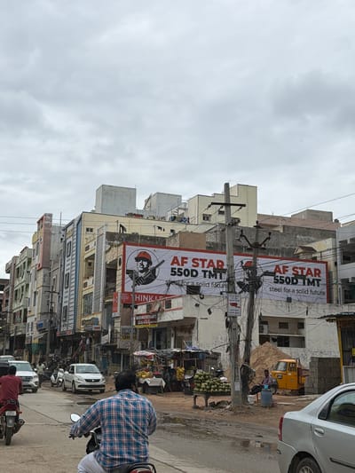 The image depicts a street scene in Shamshabad, India. In the foreground, two men are riding motorcycles, with one in a blue and white plaid shirt and white pants in the center, and another in a red shirt on the left. Several cars and motorcycles are also visible on the road. Buildings line the street, with a large billboard advertising "AF STAR 550D TMT steel for a solid future" prominently displayed on one of them. A fruit stall with piles of coconuts and bananas can be seen on the right side of the road. A yellow auto-rickshaw and a pile of sand are also present. The sky is overcast with gray clouds.