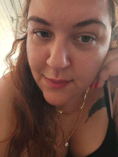 A close-up selfie of a woman with reddish-brown hair, light skin, and green eyes. She wears lipstick, and a gold chain necklace with a diamond-like pendant and a feather tattoo are visible on her chest. The background appears to be outdoors, potentially under a canopy or awning. There is no verifiable information to confirm the location to be the city of Zirndorf, Germany.