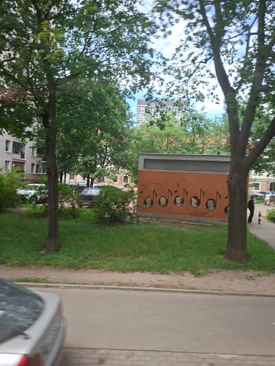 The image shows an outdoor scene with trees, buildings, and a sidewalk. In the foreground, there are several large trees with green leaves, suggesting a warm season. One tree trunk is prominent in the center. Behind the trees, there are apartment buildings with visible windows and balconies.
On an orange panel mounted on a structure, there is artwork depicting several circular portraits of people, arranged vertically and accompanied by musical notes. The portraits appear to be in a cartoonish style. Some text is visible on the orange panel, but it is not clearly legible.
To the right of the panel, there is a strip of green grass and a sandy path, leading to a paved sidewalk or road. A silver car is partially visible in the lower right corner of the image, and a dark car is parked further down the street.
In the background, there are more buildings and trees. A few people are visible walking along the sidewalk, one of whom is wearing dark clothing and appears to be looking down. The sky is partially visible and appears cloudy. The overall impression is a pleasant day in an urban environment, likely a residential area. The presence of the musical notes and portraits suggests a cultural or artistic element within the setting. The orientation of the image is sideways.