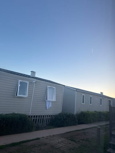 The image displays an outdoor scene in Gavà, Spain, under a clear, light blue sky that shows a subtle gradient to a yellowish hue on the right horizon and a faint aircraft contrail in the upper right quadrant. The primary subjects are a series of connected, light gray or beige prefabricated structures with dark gray shingled roofs. The leftmost visible structure features two white-framed windows; the left window is partially ajar, while the right window is fully open, pivoting outwards, with a white cloth or garment hanging from its lower frame. A small white CCTV camera is mounted on the wall between these two windows. A white chimney extends from the roof directly above. To the right, another segment of the structure continues, exhibiting multiple closed, vertical white-framed windows, and a second white chimney is visible on its roof. Dark green hedges line the base of the structures, partially obscuring a light-colored wooden lattice fence or skirting beneath the leftmost unit. A light brown paved pathway runs horizontally across the mid-ground, parallel to the structures, with dry grass and patches of earth in the immediate foreground. A wooden post and railing are partially visible on the far right foreground. No individuals or specific actions are depicted.