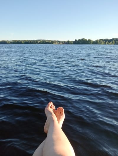 A first-person perspective shot shows a pair of bare feet and legs resting over dark blue water. The feet are crossed at the ankles, with toes pointing upwards. The skin appears to be lightly tanned and has a few small water droplets. The water is rippled, suggesting a gentle breeze or movement. In the background, a tree-lined shore is visible under a clear, pale blue sky. A few scattered buildings can be seen amongst the trees on the distant shore. The overall impression is one of relaxation by a lake. The location is identified as a lake in Suonenjoki, Finland.