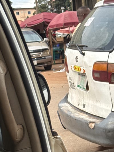 The image depicts a street scene in Orji, Nigeria, likely captured from within a vehicle. In the foreground, the interior of a car is visible on the left, with beige and black trim. Outside the car, a white van is parked, its rear end prominently featured. The van has a dented bumper and a license plate with the numbers "2395". A colorful decal with red, yellow, and white arrows is above the license plate. To the left of the van, another car, possibly a silver SUV, is partially visible. In the background, market stalls with red awnings are set up, suggesting a bustling commercial area. A person with dark skin and short hair, wearing an orange shirt, is standing near the rear of the white van, looking away from the camera. The ground is a dusty, reddish-brown surface. The overall impression is of a typical urban scene with elements of commerce and transportation.