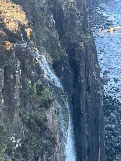 The image shows Kilt Rock, a cliff formation in Staffin, Isle of Skye, Scotland, United Kingdom. A waterfall cascades down the basalt columns of the cliff face into the sea below. The cliff consists of layered, dark grey rock formations interspersed with patches of vegetation. The sea, visible at the top, is dotted with rocks. The lighting suggests either early morning or late afternoon.