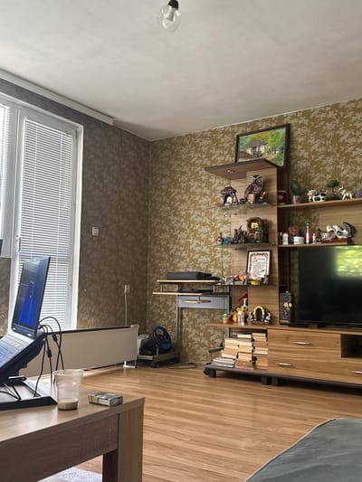 The image depicts an interior scene in Gorni Bogrov, Bulgaria. A wall-mounted wooden cabinet with shelves, a television, and various decorative objects is visible. A painting hangs on the wall next to the cabinet. A window with closed blinds is located on the left side of the image. In the foreground, a desk is present, cluttered with computer monitors, cables, a cup, and a pack of cigarettes. A gaming wheel setup is positioned next to the desk. The floor appears to be made of wood.