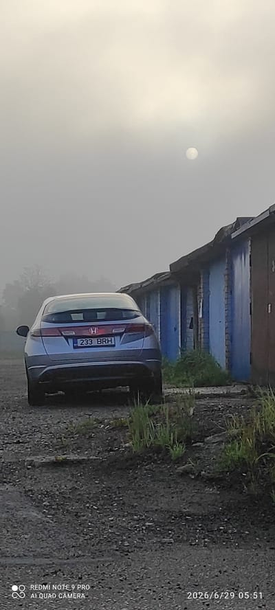 A silver Honda Civic hatchback with license plate 233 BRH is parked on a gravel surface in front of a row of blue garage doors. The sky is overcast and hazy, with a faint sun visible in the upper right quadrant. The scene appears to be in Maardu, Estonia, possibly in an area with garages or industrial buildings. There are no people or animals visible in the image. The environment is somewhat dilapidated, with weeds growing through the gravel and cracks in the garage doors. The lighting suggests it is either early morning or late evening.