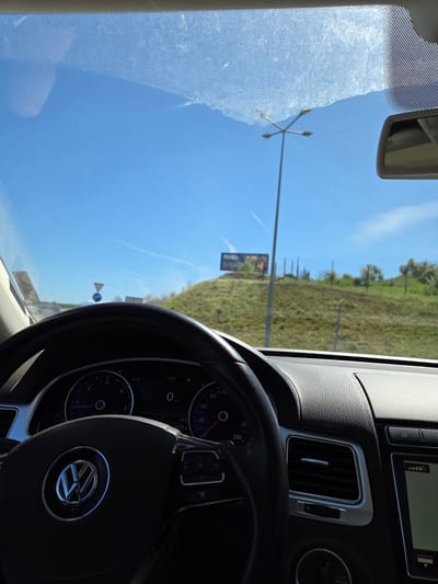The image depicts the interior of a vehicle, likely a car, with the dashboard and steering wheel prominently visible. The steering wheel features a Volkswagen logo. Through the windshield, the external environment can be observed, showcasing a green, grassy landscape under a bright blue sky. Road signs and utility poles are also visible. Based on the OCR data present, it can be confirmed that the car is located in Levunovo, Bulgaria.