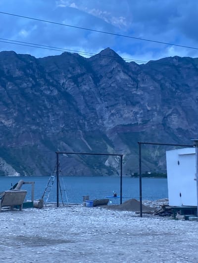 The image shows a body of water, likely a reservoir, with a steep, rocky mountain range in the background under a cloudy, dusky sky. In the foreground, there is a gravelly shore with several objects. These include a white rectangular structure with a dark vertical line on its side, a dark-colored boat, piles of sand and debris, a ladder leaning against a pole, and a bench-like structure with some dark material behind it. On the water, a person is visible in a kayak. The overall impression is of a somewhat undeveloped waterfront area at dusk or dawn, with natural elements dominating the scene. The location is identified as Irganay, Russia.