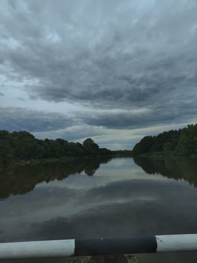 This is a full-frame, eye-level, medium shot of a wide, calm river. The river dominates the lower half of the frame, reflecting the dark, cloudy sky and the dense green treeline that borders its banks. The sky is filled with heavy, gray storm clouds, creating a somber atmosphere. The treeline stretches across the horizon, with lush foliage visible on both sides of the river. The foreground of the image features the top of a metal railing, painted white and black, which is partially obscuring the lower edge of the scene. The overall impression is one of natural beauty under an impending storm. The location is identified as the river in Tumutuk, Russia.